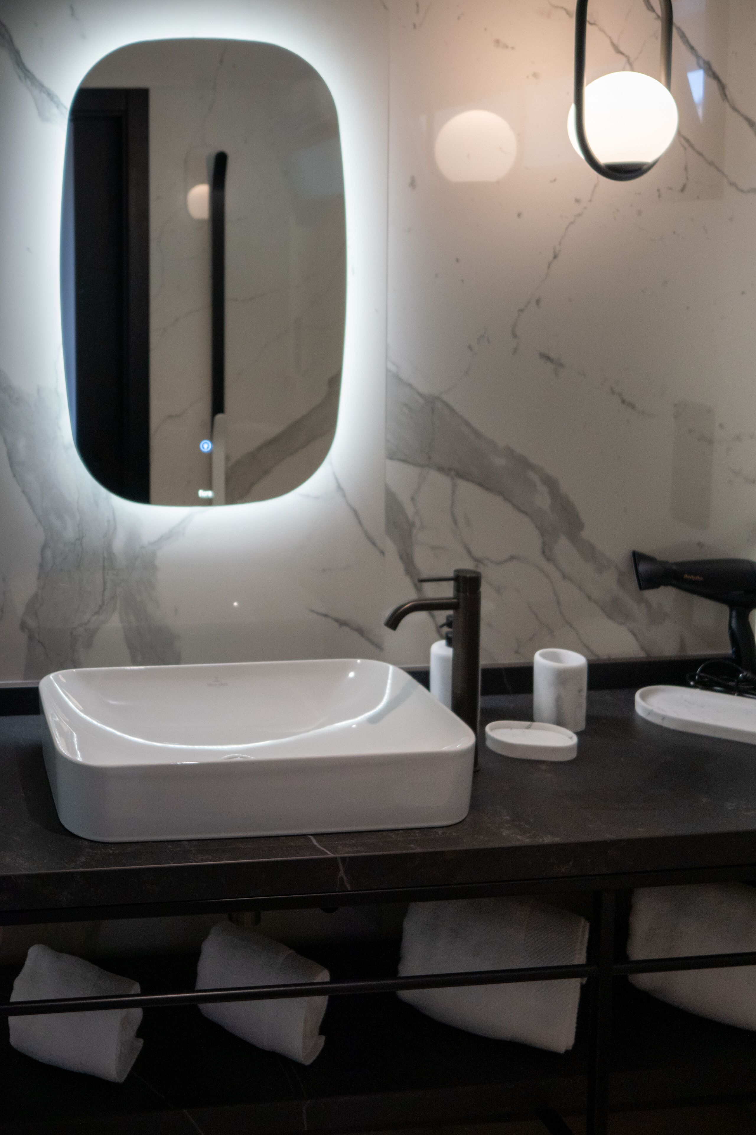 Un baño moderno con un lavamanos ovalado grande, un espejo, un secador de pelo y un perchero para toallas.