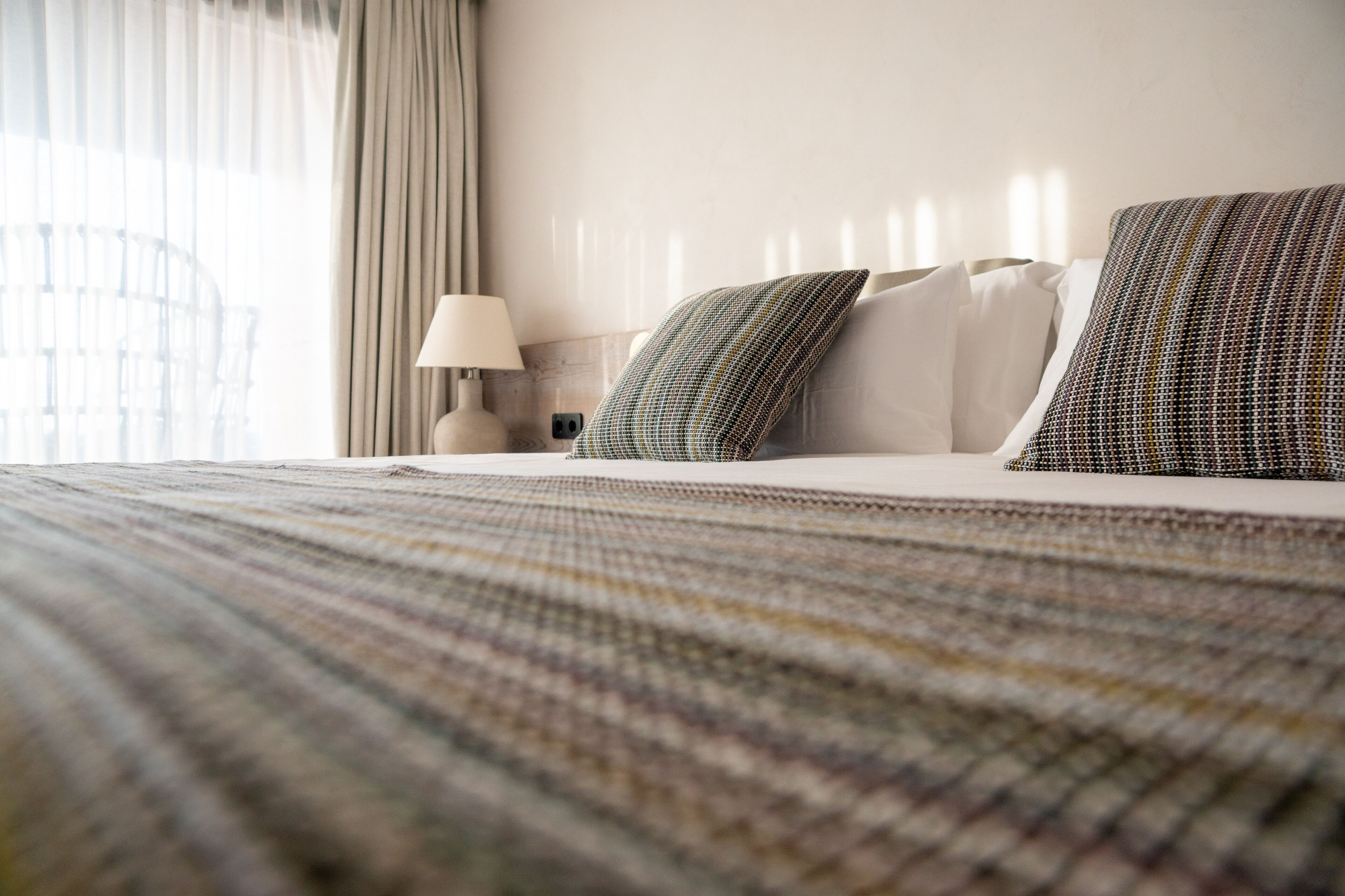 A neatly made bed with patterned pillows, a lamp, and a view of a wicker chair through sheer curtains.
