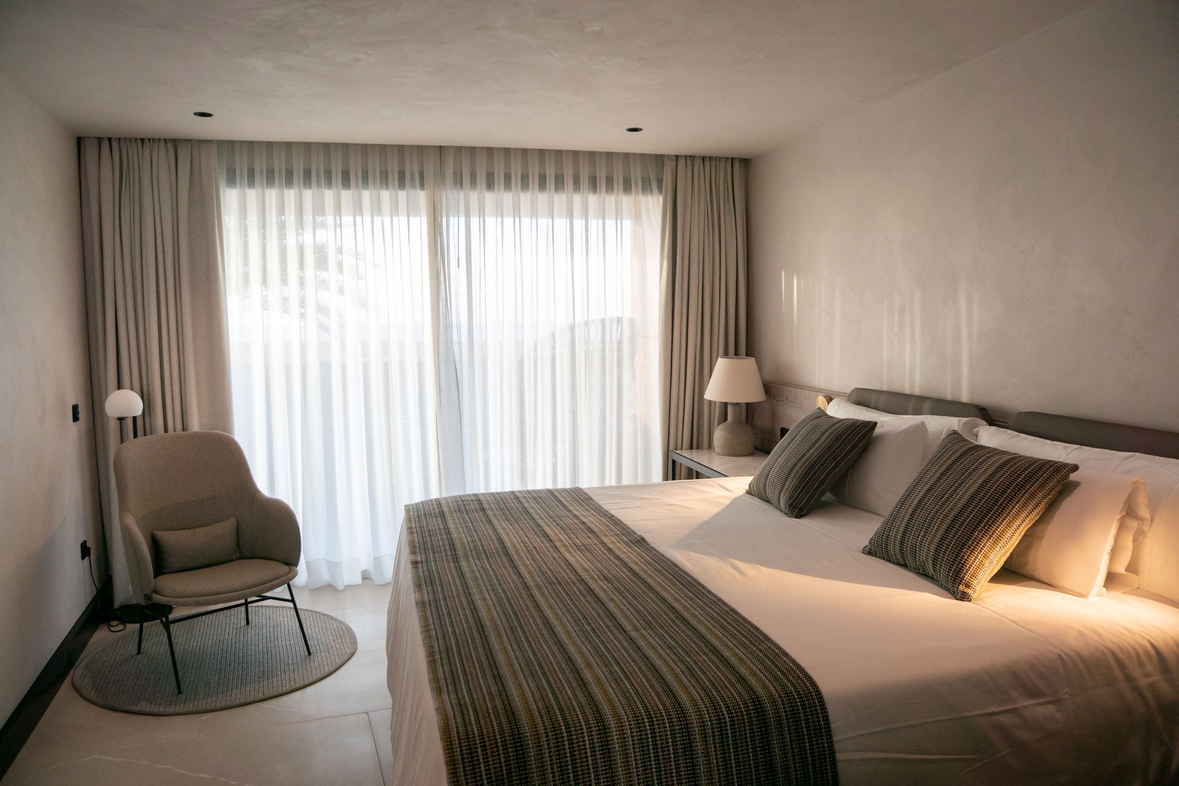 A hotel room with a large bed, a chair, a lamp, and sheer curtains.