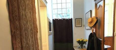 A narrow hallway with a bench, a hanging light fixture, a window with a curtain, and a door with a hat on a stand.