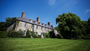 A large, historic stone building with multiple chimneys, surrounded by a well-maintained lawn and dense trees.