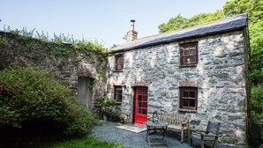 A stone building with a red door, a small garden with a table and chairs, and a chimney.