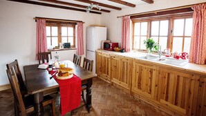 A cozy kitchen with wooden cabinets, a dining table with chairs, and a red tablecloth. There is a refrigerator, a microwave, and a sink with a window above it. - Erw Rhys - sleeps 6 guests  in 3 bedrooms (Holyhead)
