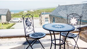 Eine Terrasse mit einem runden Tisch und zwei Stühlen, mit Blick auf die Küste, mit Steinbauten und einem geparkten Auto.