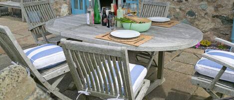 Een rustieke buiten eethoek met een ronde houten tafel, vier houten stoelen met blauwe kussens en een barbecue met worsten.
