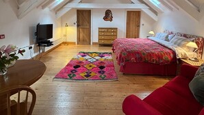 Une chambre à coucher aménagée dans un grenier, avec un plafond en pente, des poutres en bois, un lit recouvert d’une courtepointe rouge, une commode en bois, un téléviseur et un tapis aux couleurs vives.