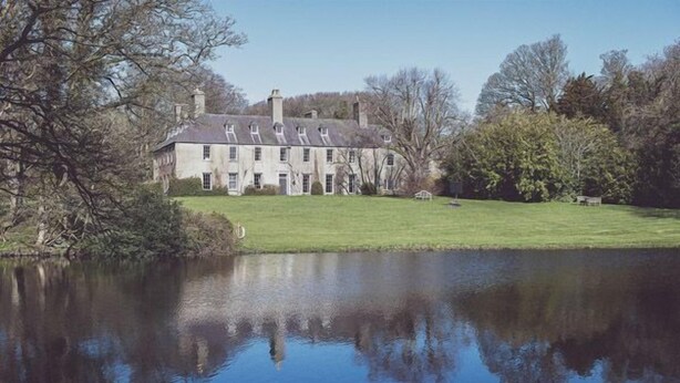 Un grand bâtiment historique, doté de nombreuses cheminées, situé au bord d’un lac paisible, entouré d’arbres et d’une pelouse bien entretenue.