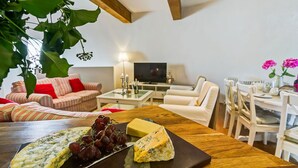 Un salon chaleureux avec une table en bois dressée pour déguster du fromage et des raisins, un canapé et un téléviseur en arrière-plan.
