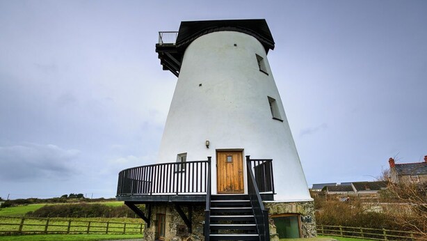Un moulin à vent blanc, doté d’une porte en bois et de balustrades noires, entouré d’un chemin de gravier et de verdure.