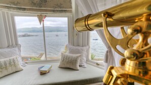 Una habitación con un amplio ventanal con vista a un cuerpo de agua, un telescopio y un libro en el alféizar.