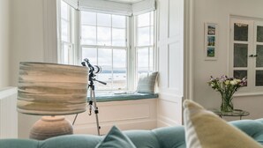 Et værelse med karnap der vender ud mod en strand, et teleskop på stativ og en vase med blomster på et sengebord.