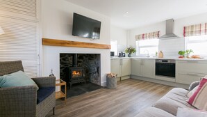 A cozy living room with a fireplace, a television mounted above it, and a kitchen with a window. - Pandy Aberffraw -  a dog that sleeps 4 guests  in 2 bedrooms (Ty Croes)