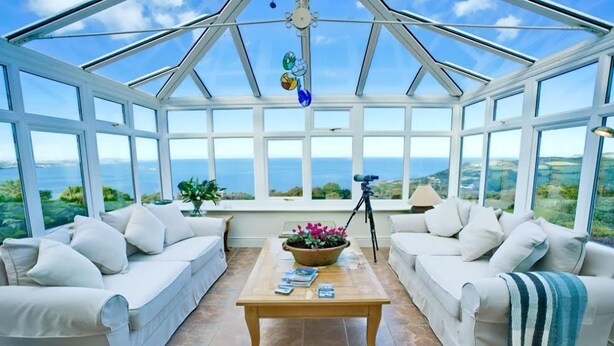 A conservatory with a clear view of the sea, furnished with white sofas and a wooden table. - Tan Y Bryn -  a sea view that sleeps 10 guests  in 5 bedrooms (Llanddona)