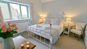 A bedroom with a bed, bedside tables, a chair, a window with curtains, and a vase with flowers. - Afon Menai -  a hot tub that sleeps 4 guests  in 2 bedrooms (Llanfairpwllgwyngyll)