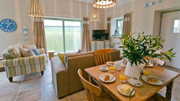 A well-lit living room with a sofa, dining table, and a TV. There is a large window with curtains, a clock, and a plant. - Afon Menai -  a hot tub that sleeps 4 guests  in 2 bedrooms (Llanfairpwllgwyngyll)