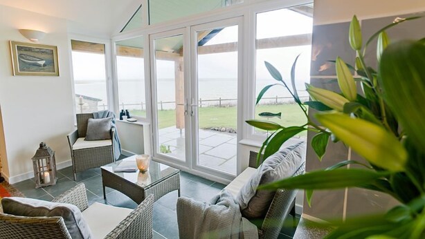 Ein geräumiges Zimmer mit einer großen Schiebeglastür, die auf eine Terrasse mit Meerblick führt. Es befinden sich Korbmöbel und ein Blumentopf dort.