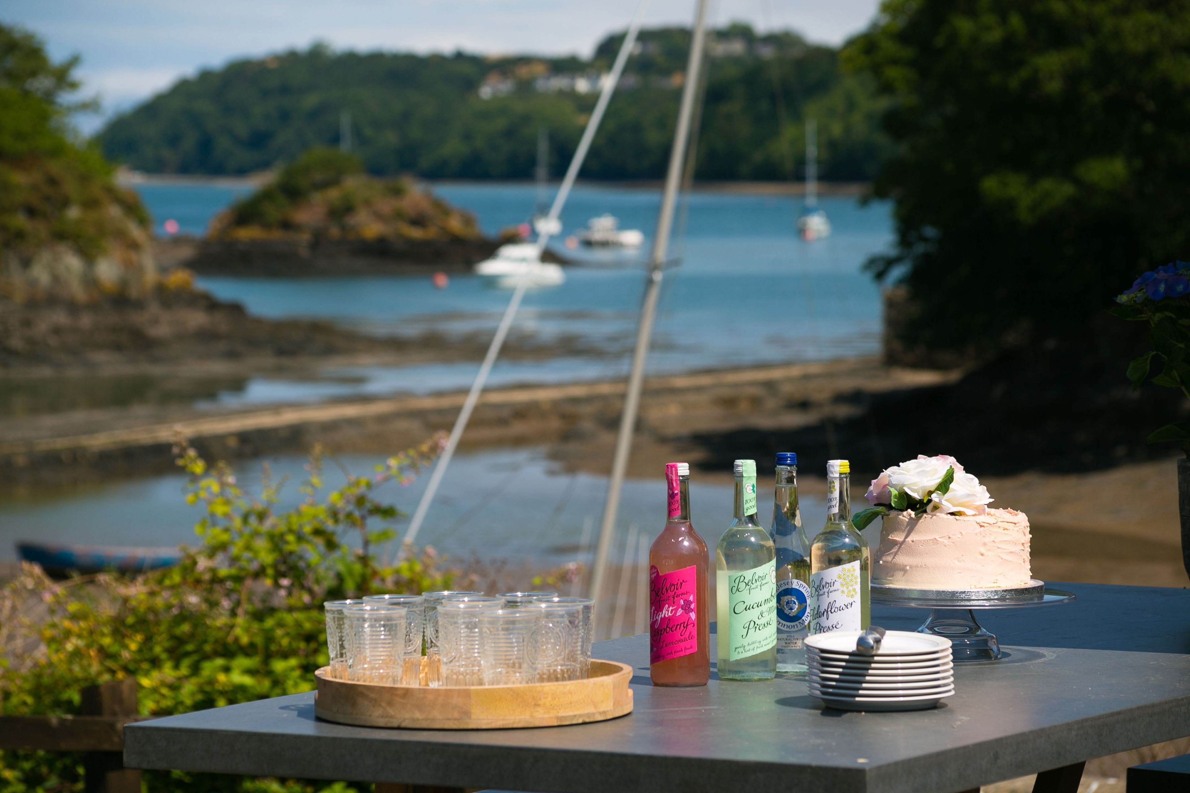 A table with bottles, glasses, and a cake, overlooking a body of water with boats.