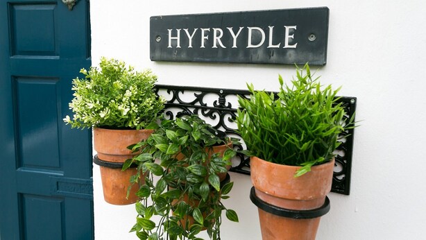 Un letrero que dice "HYFRYDE" sobre una pared blanca, con dos macetas de terracota con plantas verdes a cada lado.