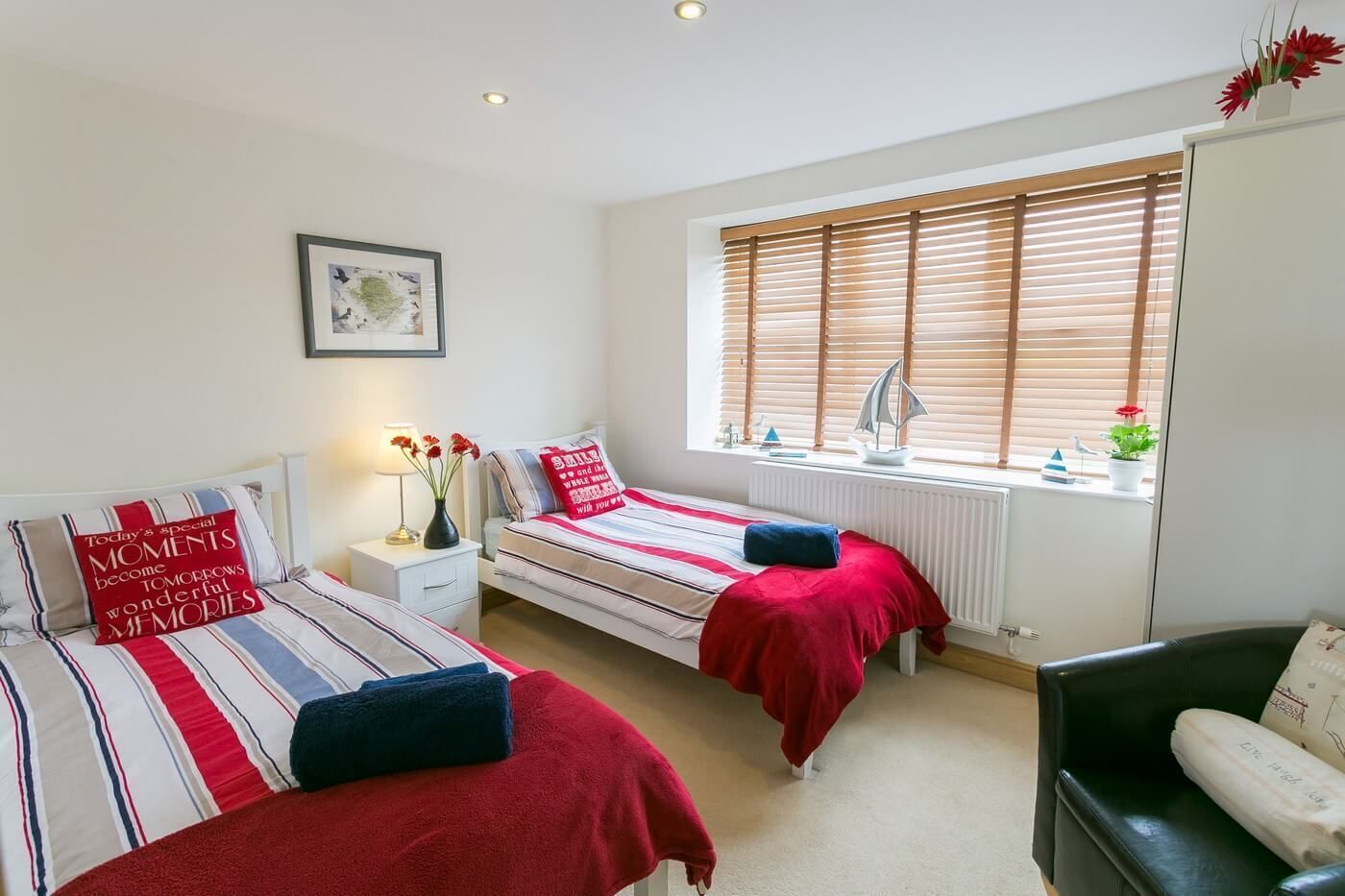 A bedroom with two beds, a window with blinds, a radiator, a desk with a lamp, and a chair.