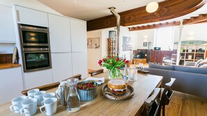 Una cocina moderna con hornos empotrados, un comedor con mesa de madera dispuesta para una comida, y una sala de estar acogedora con sofá y chimenea.