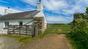 A rustic stone house with a white exterior, a wooden gate, and a dirt path leading to a green field.