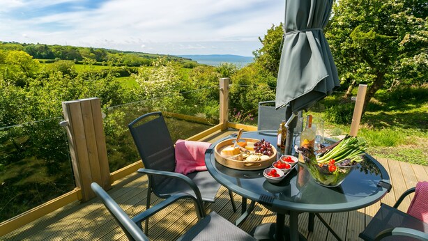 An outdoor dining area with a table set for two, surrounded by chairs, a wooden deck, and a view of a lush landscape.