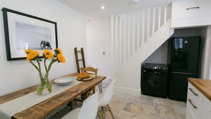 A compact kitchen with a washing machine, a wooden dining table with chairs, and a framed picture on the wall. - Shell Cottage -  a dog that sleeps 4 guests  in 2 bedrooms (Menai Bridge)