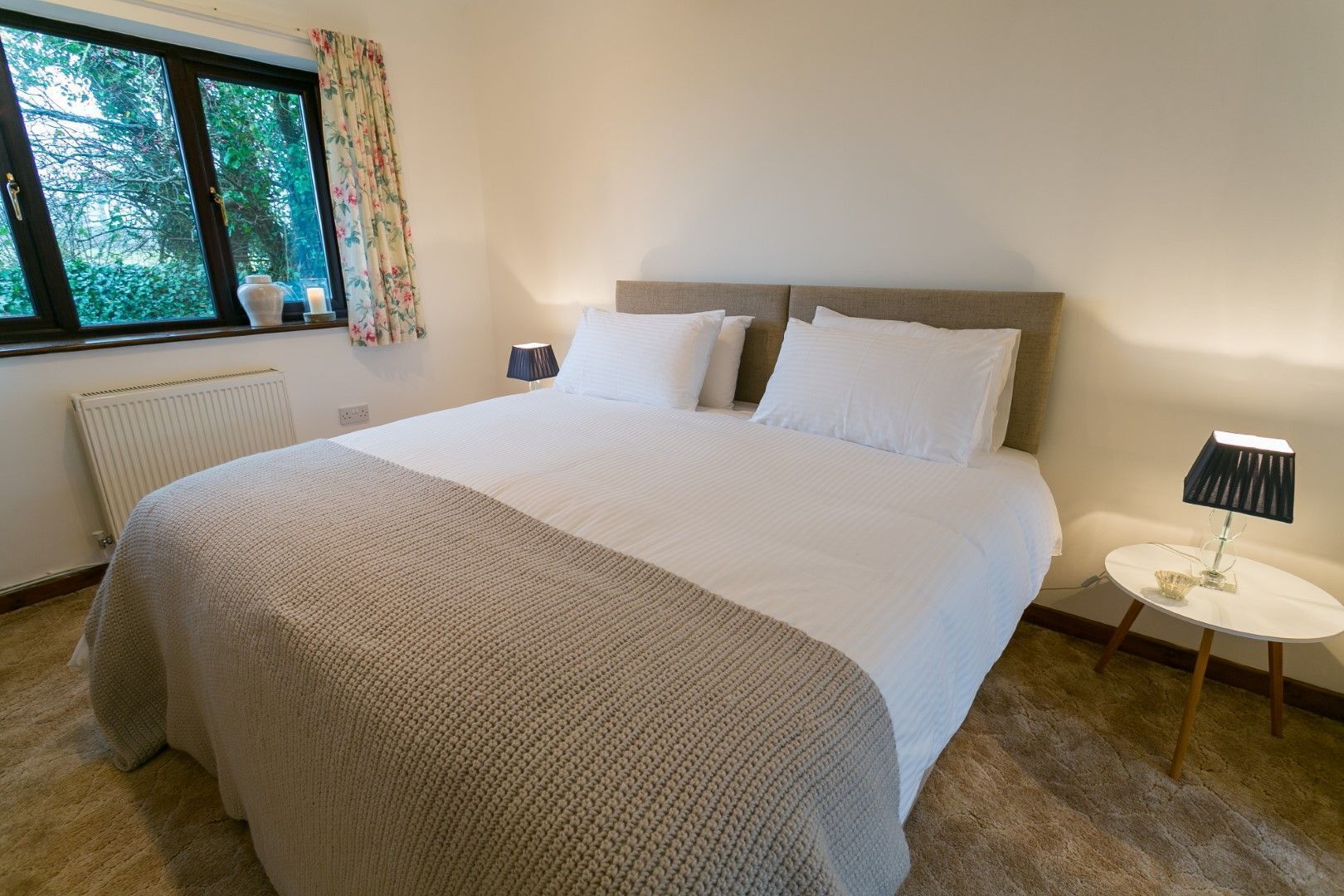 A neatly made bed with a beige blanket, a bedside table with a lamp, and a window with curtains.