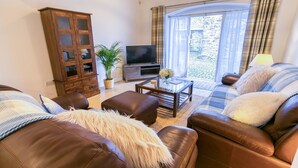 A living room with brown leather sofas, a glass coffee table, a wooden cabinet, a television, and a view through a window with curtains.
