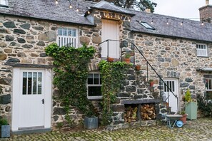 A stone building with a white door, windows, and a small porch area with a firewood stack and potted plants.