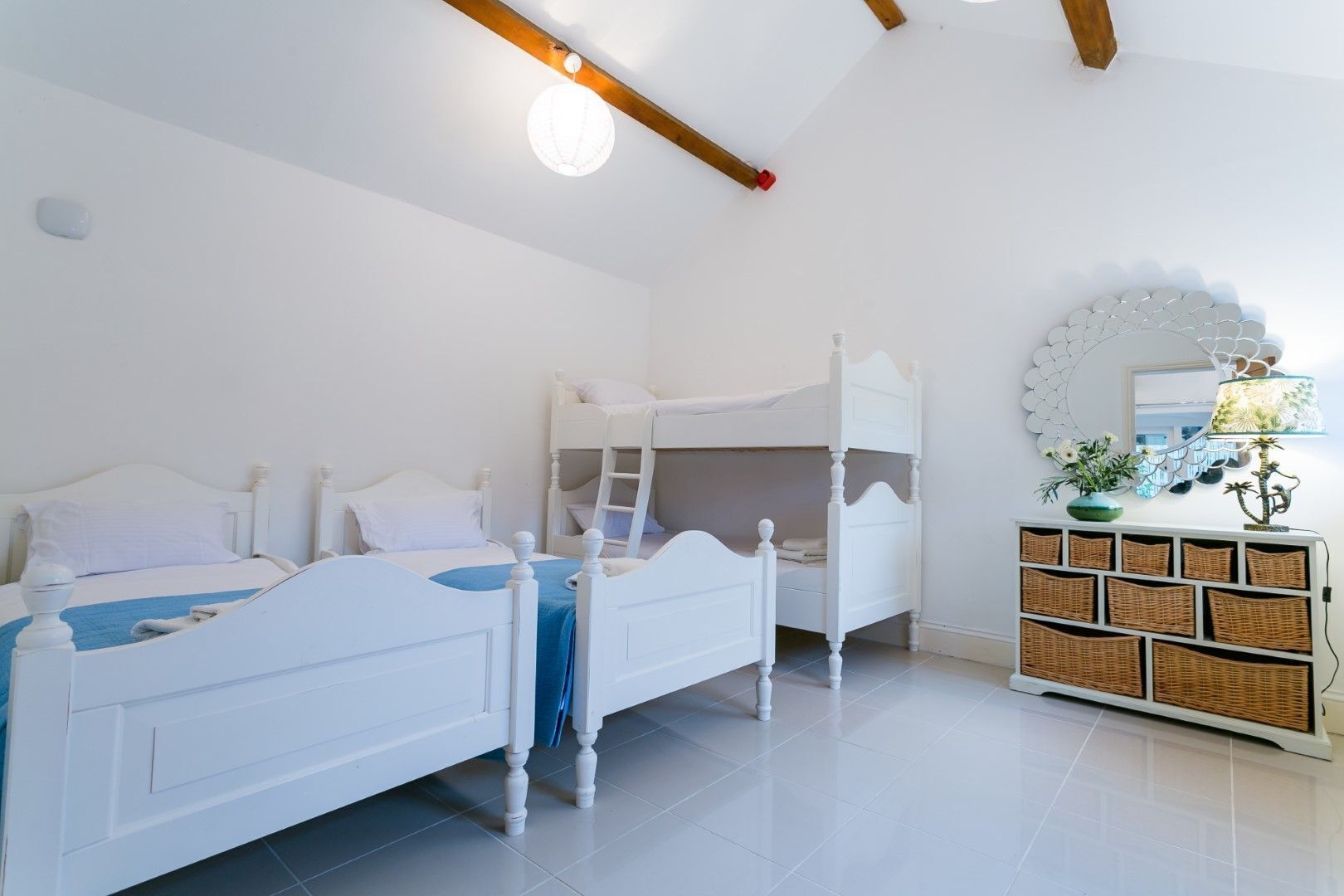 Une chambre avec deux lits blancs, un lit superposé, un miroir, des paniers en osier et un luminaire au plafond.