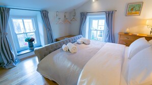 Ein helles Schlafzimmer mit einem großen Bett, zwei Fenstern mit Vorhängen, einem Nachttisch mit Lampe und einem Bild an der Wand.