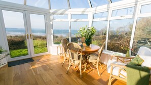 A conservatory with a wooden floor, wicker chairs, a round table, and a view of the sea.