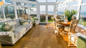 A conservatory with a wooden floor, a sofa with patterned cushions, wicker chairs, and a table with a vase of flowers.