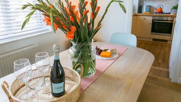 A dining table with a bottle of champagne, glasses, a vase with flowers, and a plate with cheese and grapes. - Sea Bell -  a coastal that sleeps 6 guests  in 3 bedrooms (Holyhead)