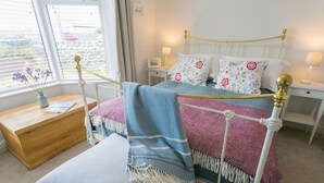 A neatly arranged bedroom with a metal-framed bed, a wooden bedside table, a small bedside table with a lamp, and a window with blinds. - Sea Bell -  a coastal that sleeps 6 guests  in 3 bedrooms (Holyhead)
