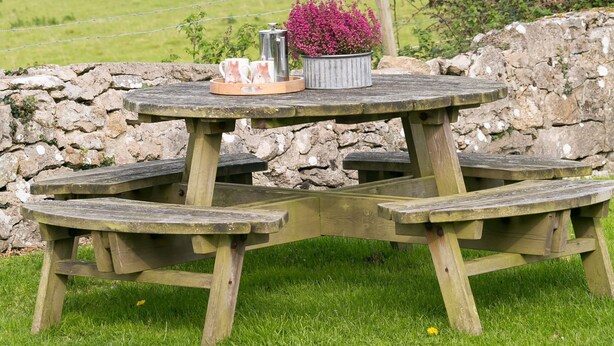 A wooden picnic table with a metal pot of flowers and a teapot, set against a stone wall with a view of a coastal landscape. - Tyn Lon -  a sea view that sleeps 9 guests  in 5 bedrooms (Moelfre)