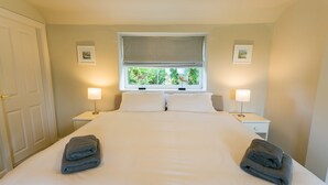 A neatly made bed with white linens and two folded towels on each side. There are two bedside lamps, a window with a view of greenery, and a door to the left. - Tyn Lon -  a sea view that sleeps 9 guests  in 5 bedrooms (Moelfre)