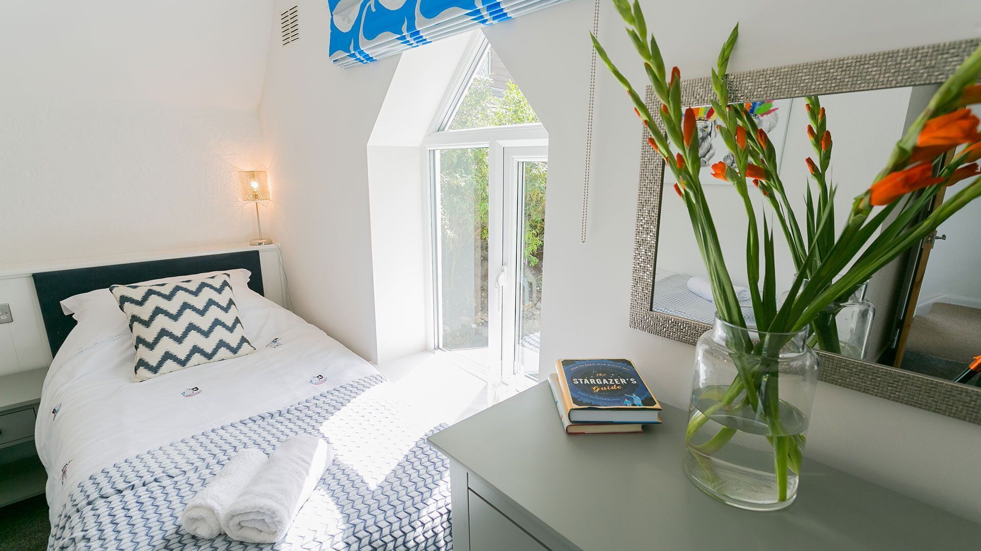 A bedroom with a bed, a bedside table with a book, a mirror, and a window with a view of greenery.