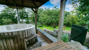 A wooden hot tub under a covered area with a stone wall and a wooden deck.