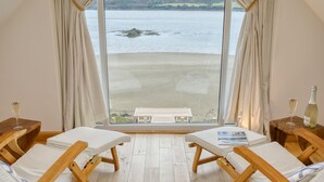 A room with two wooden lounge chairs, a wooden table, and a view of a beach through large windows. - House on the Beach -  an on the beach that sleeps 9 guests  in 4 bedrooms (Holyhead)