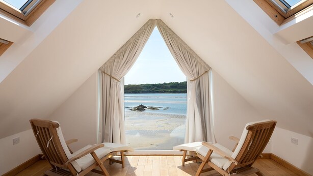 A room with a sloped ceiling, two wooden chairs facing a beach view through a window with curtains. - House on the Beach -  an on the beach that sleeps 9 guests  in 4 bedrooms (Holyhead)