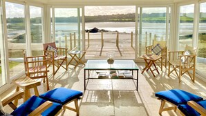A sunlit room with large windows, wooden chairs, a table with books, and a view of a beach. - House on the Beach -  an on the beach that sleeps 9 guests  in 4 bedrooms (Holyhead)
