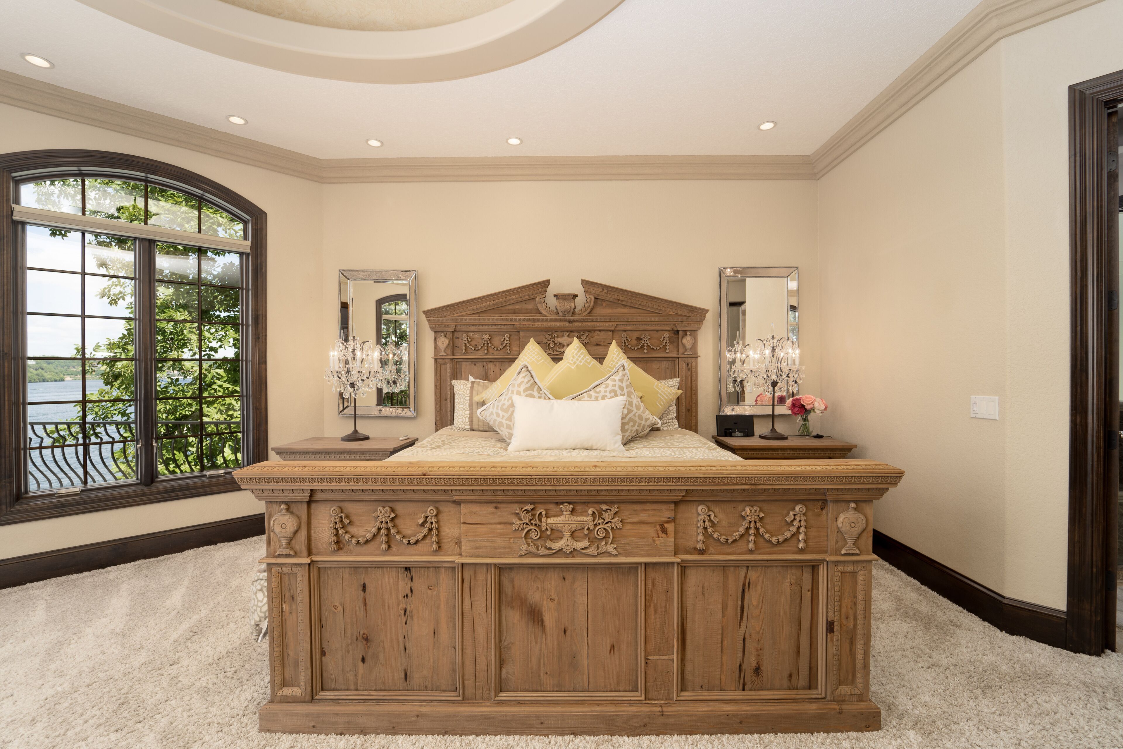 A large bed with ornate headboard, two bedside tables with lamps, and a large window with a view of greenery.