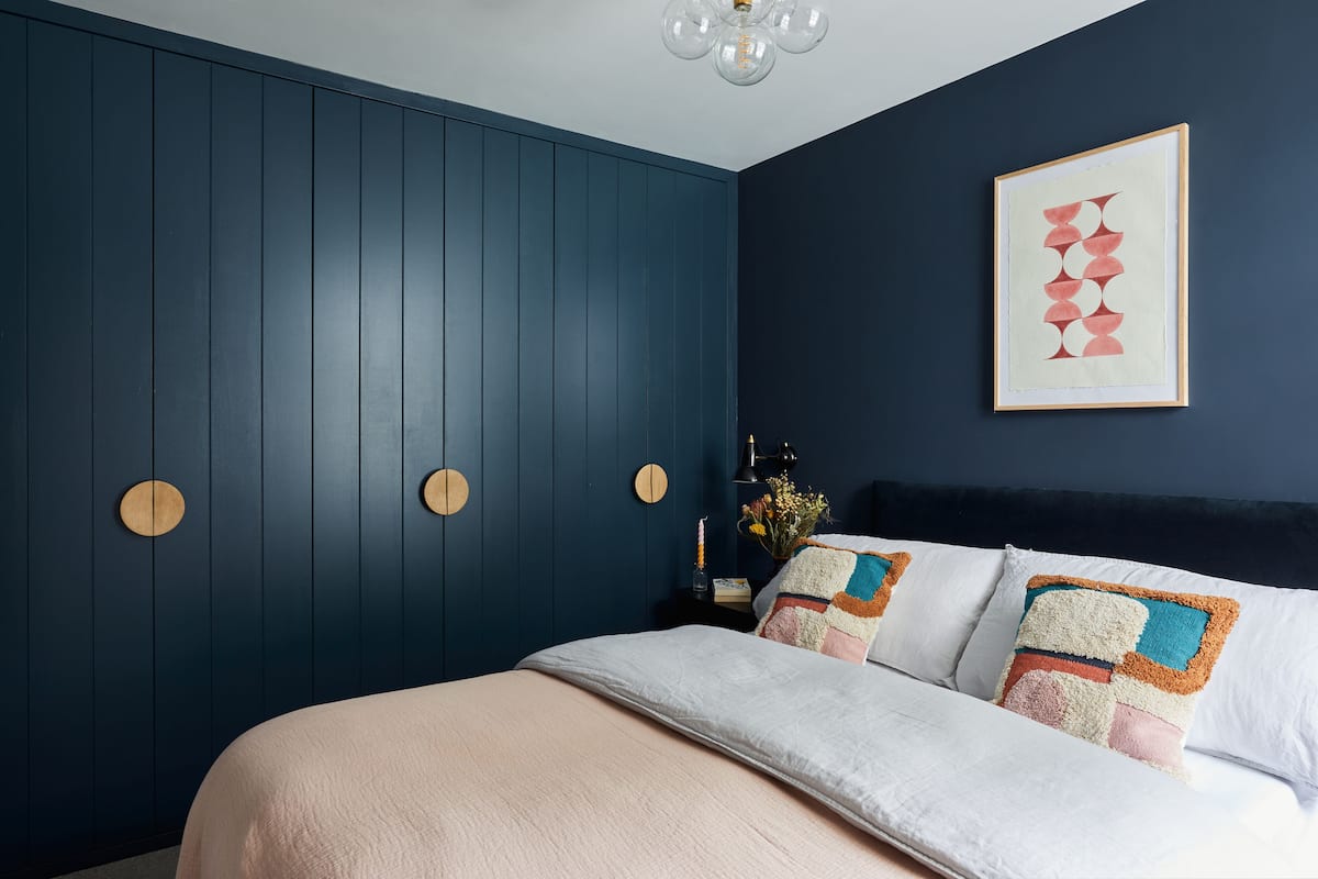 A bedroom with a bed, a dark blue wall with circular cutouts, a framed artwork, and a nightstand with a candle and flowers.
