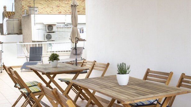Un conjunto de mesa y sillas de madera en un balcón con plantas en macetas, vista a edificios y unidades de aire acondicionado.