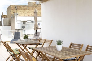 A wooden table and chairs set on a balcony with potted plants, a view of buildings, and air conditioning units.