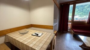 Een kleine, gezellige kamer met een houten bankje, een tafel met een geruit tafelkleed, een afstandsbediening en een raam met uitzicht op een bebost gebied.