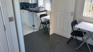 Een kleine keuken met witte kasten, een klein aanrecht en een raam met gordijnen. Er is een witte deur en een tafel met twee stoelen.