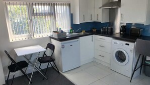 Een compacte keuken met een wasmachine, een eettafeltje met stoelen en een raam met zonwering.